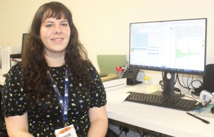 Maite Devos Lozano is sitting at a desk in front of a computer screen and keyboard. She is wearing a black dress with a flowery pattern and has long brown hair.