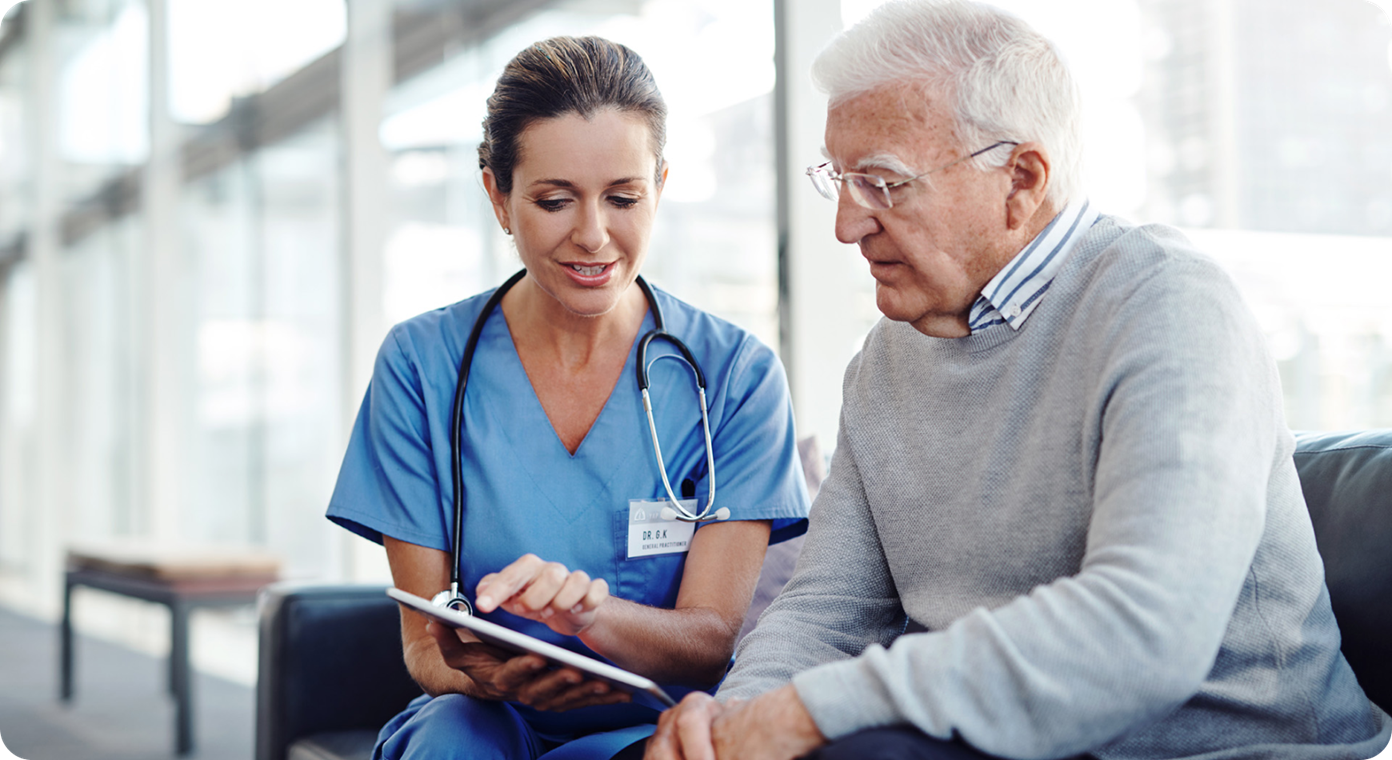 A female medical professional is sitting beside an older man. She is pointing to and showing the man something on a tablet.