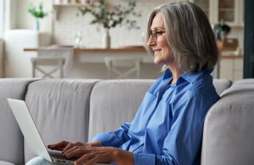 A woman is sitting on a sofa in a home setting and is typing on a laptop.