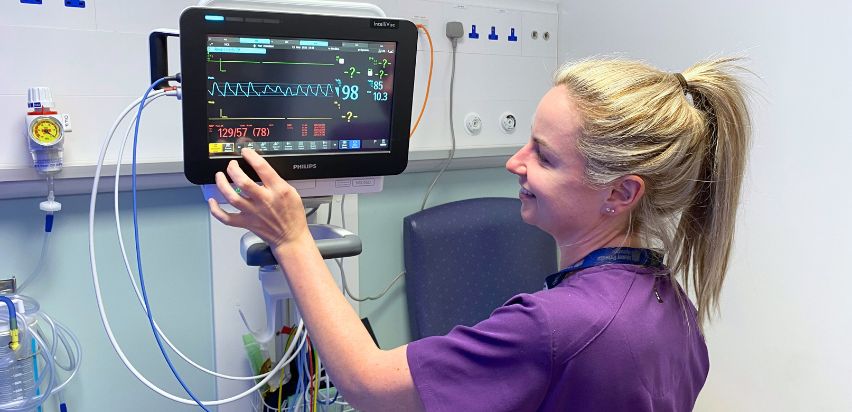 A blonde nurse, wearing a purple uniform, looking at a heart monitor screen